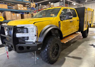 Yellow work/service truck in a warehouse-like shop, with a heavy-duty front bumper and a large utility rear body, on a workshop floor.
