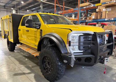 Yellow utility service truck with heavy-duty front bumper and winch in a warehouse workshop area.