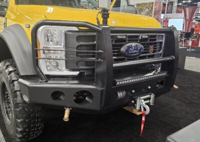 Front-left view of a bright yellow Ford pickup with a heavy-duty black off-road bumper and winch at an auto show, showing a mounted device on the hood rail.