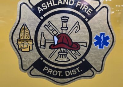 Ashland Fire Protection District badge with a red firefighter helmet and tools on a yellow surface.