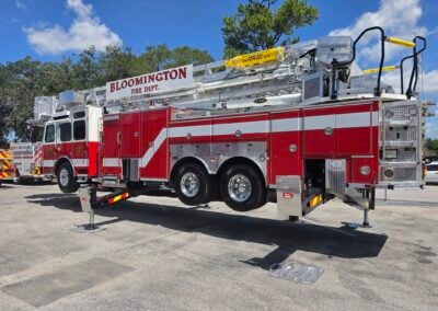 Red fire engine with an extended aerial ladder and Bloomington Fire Department sign, parked on a paved lot under a blue sky.