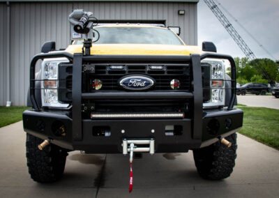 Front view of a yellow Ford off-road truck with a heavy bull bar, LED lights, and a red tow strap hanging from the bumper.