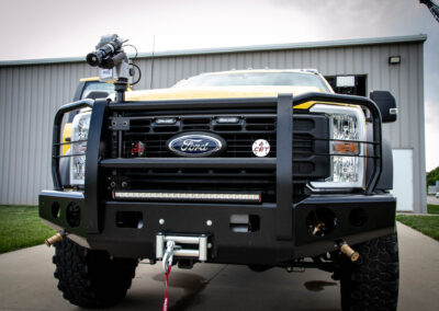 Front view of a yellow Ford monster truck with a heavy grille guard, winch, and LED light bar.