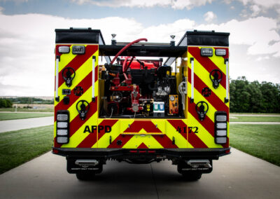 Rear view of a bright yellow-and-red fire engine with an open equipment panel, red hoses, and gauges visible at the back.