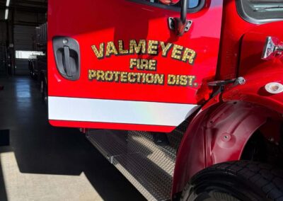 Red fire truck cab with bold 'Valmeyer Fire Protection Dist.' lettering, chrome side mirror, and front wheel, inside a fire station.