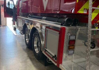 Fire rescue truck inside a station bay, side view with open door and shiny diamond-plate panels and hoses along the chassis.