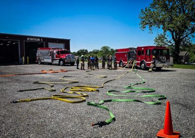 Firefighters stand in turnout gear beside two red fire engines outside Station 1, with hoses laid out on a gravel lot.