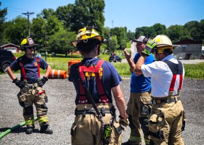 Group of firefighters in turnout gear and helmets standing on gravel, discussing a yellow hose outdoors on a sunny day.