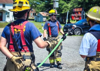 Firefighters in full turnout gear and yellow helmets handling a green fire hose on a gravel lot in a residential area.