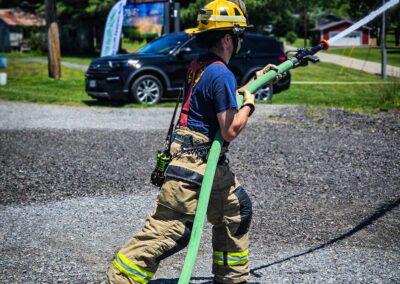Firefighter in turnout gear aiming a large green hose and spraying water on a gravel lot with trees in the background.