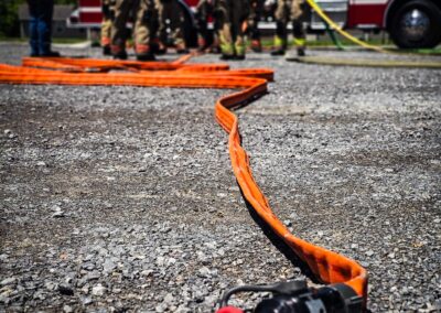 Firefighters in gear stand near a red fire truck with orange hoses laid out on gravel in the foreground.