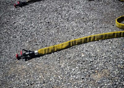 Yellow and gray fire hoses laid out on a rough gravel surface outdoors, as if at a training site or scene.