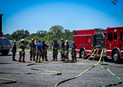 Group of firefighters in turnout gear standing beside a red fire truck with coiled hoses on a gravel lot.