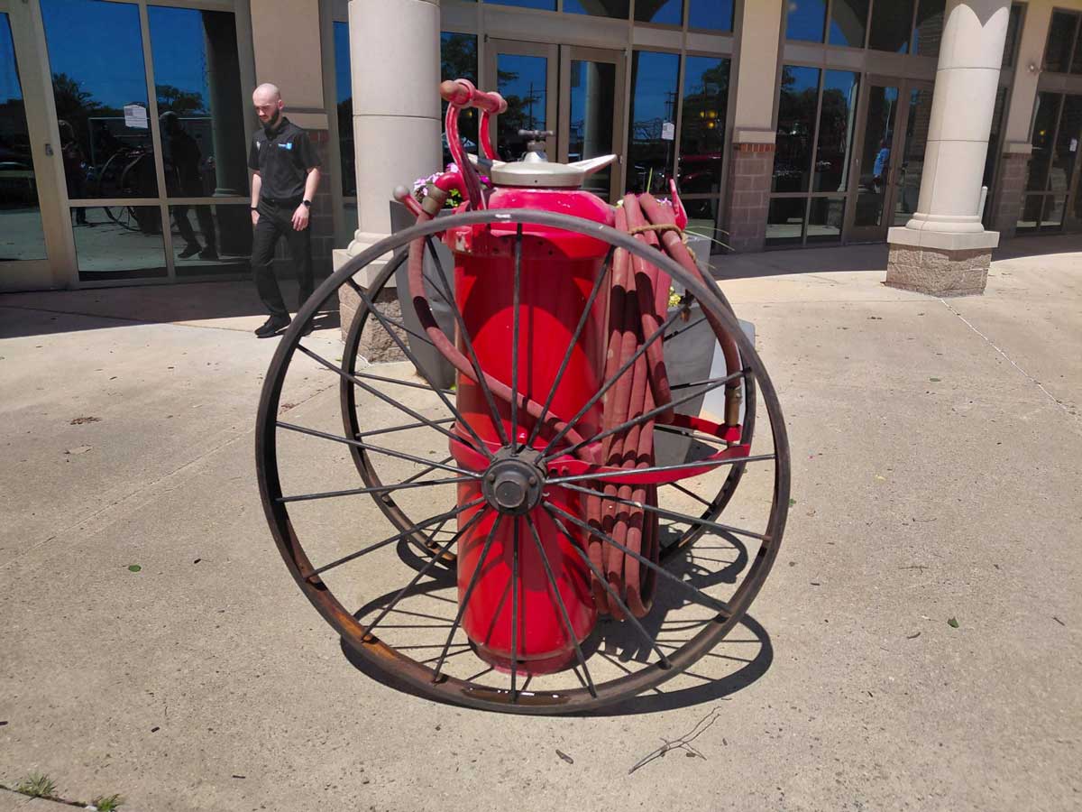 Red vintage fire hose cart with a long coiled hose outside a glass-front building; a man in black walks nearby.