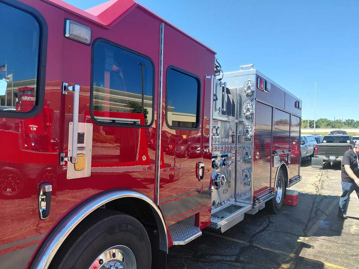 Red fire truck parked in a parking lot, chrome compartments and gauges along its side, a person standing nearby under a clear blue sky