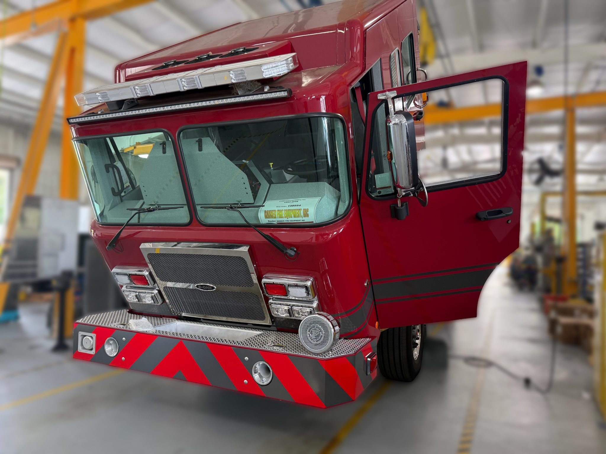 Front-left view of a red fire engine inside a workshop, with the cab door open and chevron-patterned rear bumper visible.