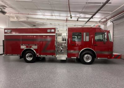 Red fire engine parked indoors in a clean, well-lit fire station bay, showing multiple equipment compartments and chrome wheels.