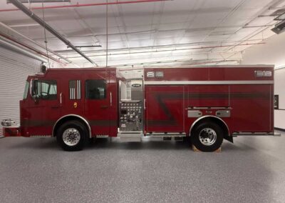 Red fire engine parked indoors in a station bay, showing cockpit cab and attached rear apparatus compartments.