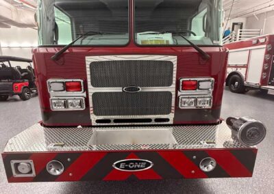 Front view of a red fire truck inside a fire station, chrome grille, chevron-pattern bumper with E-ONE logo and emergency lights on top