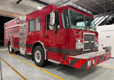 Red fire engine parked inside a fire station bay with chrome accents and yellow floor lines, facing right.”] ,