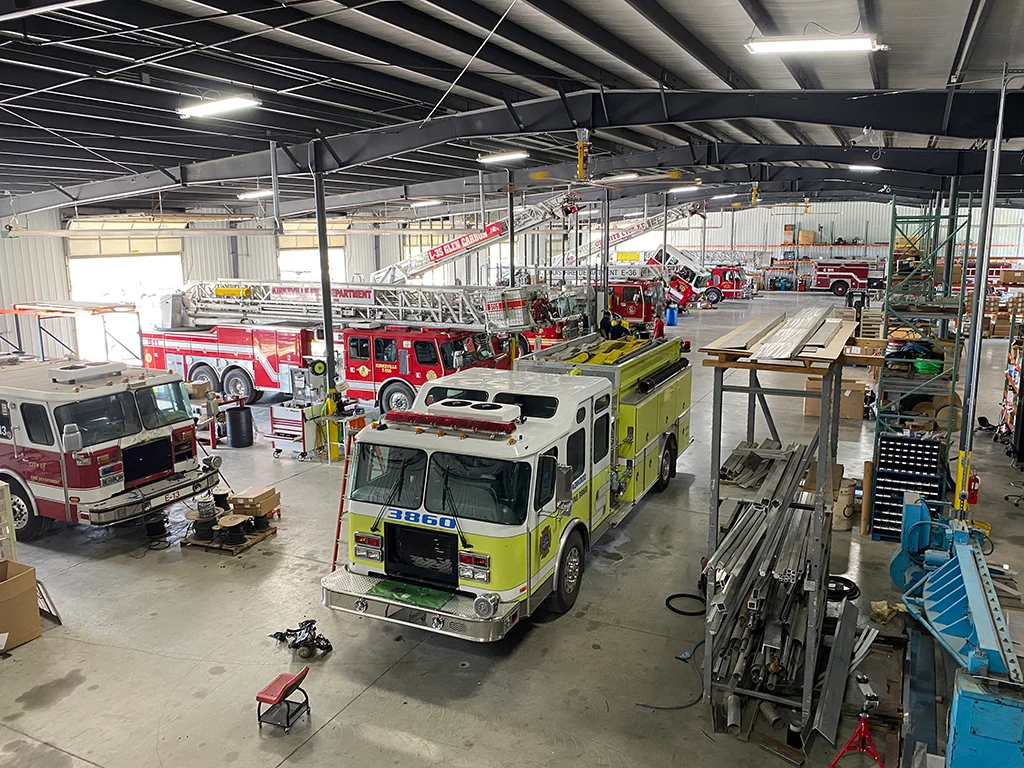 Inside a large fire station maintenance bay with lime-green pumper in the foreground and red ladder trucks in the background, plus equipment and tools around.