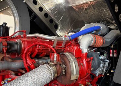 Close-up of a red industrial engine with a braided exhaust hose and blue intercooler hose in a metal compartment lined with reflective insulation panels.