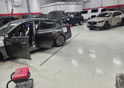 Indoor auto body shop with multiple cars being serviced; a gray sedan has its doors open and a damaged white SUV is visible in the background near an aluminum repair station sign.