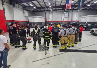 Group of firefighters in turnout gear standing in a circle inside a large training facility, with vehicles and flags overhead.