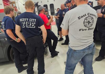 Group of firefighters in uniform gather in a workshop around a gray car, listening to a briefing.
