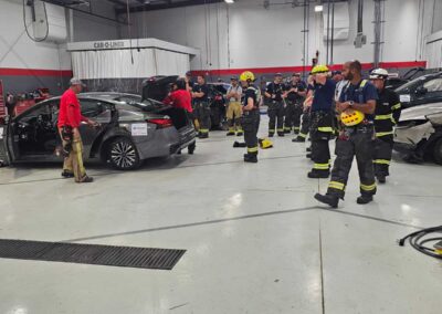 Firefighters in turnout gear gathered around a damaged car inside a large training facility, American flag overhead6