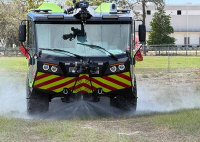 Front view of a fire rescue truck with high-visibility chevrons, spraying water on grass in a parking lot area