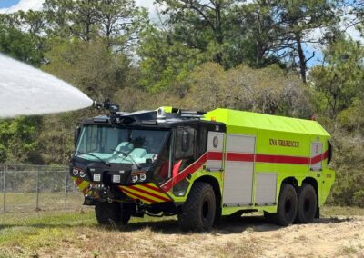 Bright yellow fire rescue truck spraying water in a brushy outdoor area with trees behind it.