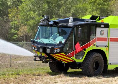 Bright green fire truck with water cannon spraying a jet of water toward the ground, outdoors near a chain-link fence.
