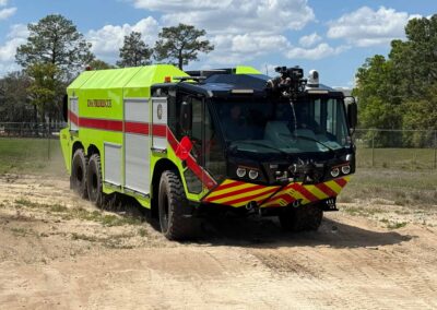 Bright lime-yellow fire rescue truck parked on a dirt field, with red stripes and equipment compartments visible under a blue sky.
