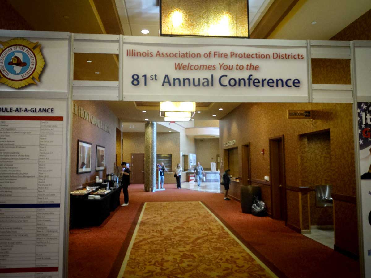 Conference hallway with a banner: 'Illinois Association of Fire Protection Districts Welcomes You to the 81st Annual Conference'; people near a refreshment table in the background.