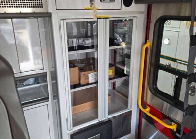 Interior of an ambulance with a glass-front cabinet containing boxes on shelves, a small control panel above, and a yellow handrail beside a window.