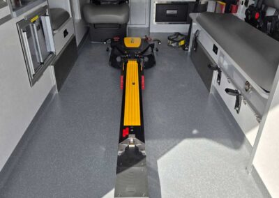 Interior of an ambulance with a wheeled patient stretcher centered on a yellow track, seating and medical gear along the sides.