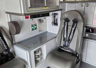 Interior of an ambulance with grey seated EMS chair and black safety belts, medical cabinets, and control panel nearby.