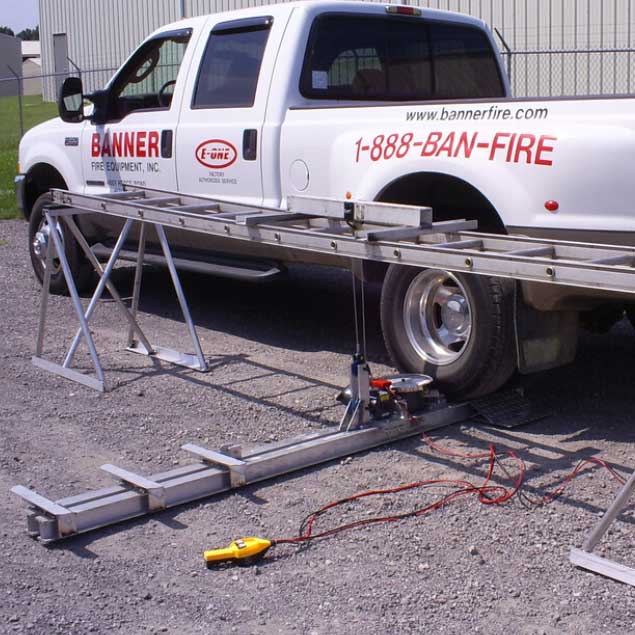 A worker in a black shirt attaches a small tag to a cable on a metal trailer frame, with a service truck and yellow tool bin in the background.