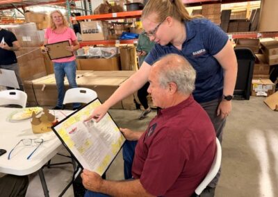 Volunteer helps an older man read a laminated form at a table during a warehouse charity event; other volunteers stand nearby.