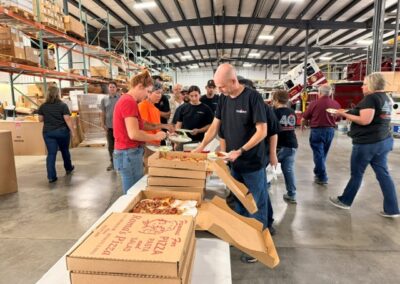 Group of volunteers in a large warehouse serving fresh pizza from open pizza boxes laid out on a table; people line up to grab slices while others sort boxes and packages on shelves in the background.