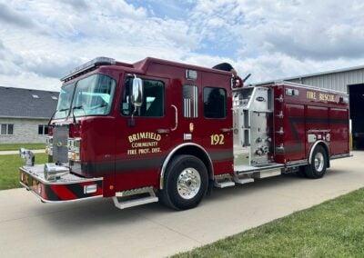 Red fire engine parked on a driveway with a firefighter on the roof handling equipment, Brimfield Community Fire Prot. Dist. label visible and an American flag graphic on the door.