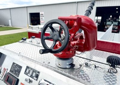 Close-up of a red fire-pighting pump handle with a black wheel on a diamond-plate control panel, gauges and labels nearby.