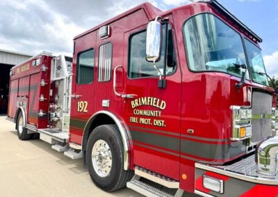 Red fire engine with 'BRIMFIELD COMMUNITY FIRE PROT. DIST.' lettering, chrome mirrors, and metal steps parked outdoors at a station bay.