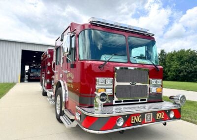 Red fire engine parked at a station bay with a chrome grille and emergency lights on the roof, front view outside on a sunny day.