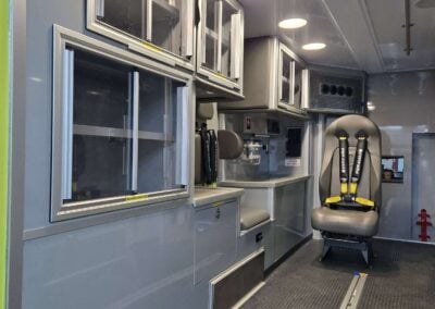 Interior of an ambulance showing gray cabinets with glass doors, storage drawers, and a padded patient seat with yellow restraints off to the side (medical vehicle).