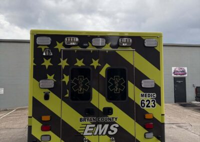 Rear of a bright yellow-and-black EMS ambulance with star pattern and two rear doors, lights, and 'Bryan County EMS' lettering visible against a cloudy sky.