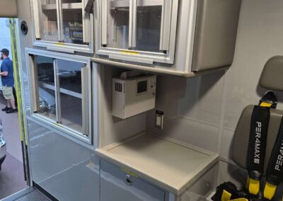 Interior of an ambulance showing wall-mounted metal storage cabinets with glass doors, a small countertop, and a grey seat with yellow safety straps nearby