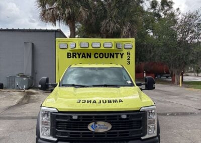 Front view of a bright yellow Bryan County ambulance parked in a lot with palm trees and cloudy sky behind it. Educational and informative image for emergency services context.
