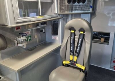 Interior of an ambulance with a padded seat and yellow-buckled restraints, medical equipment cabinets, and a ventilator intake on the wall behind.